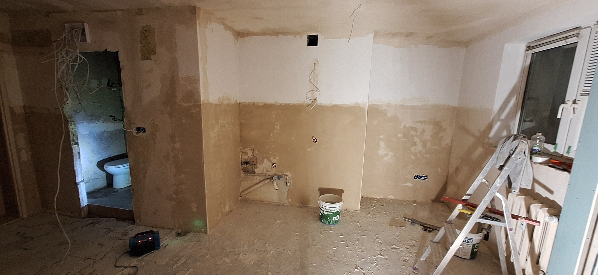 Wnętrze w trakcie prac wykończeniowych: częściowo wyszpachlowane ściany, widoczne instalacje, drabina oparta o okno, otwarte przejście do łazienki z widoczną toaletą.