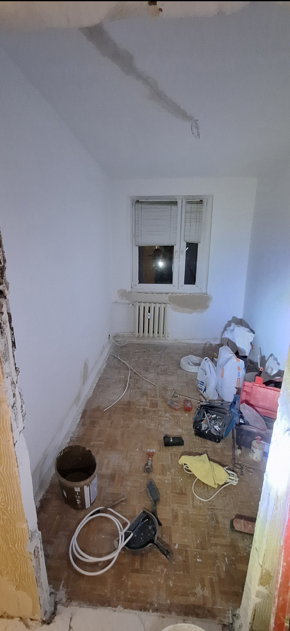 Pokój w trakcie remontu: ściany po szpachlowaniu, widoczne narzędzia, wiadro z farbą i worki z materiałami budowlanymi na podłodze z parkietu.