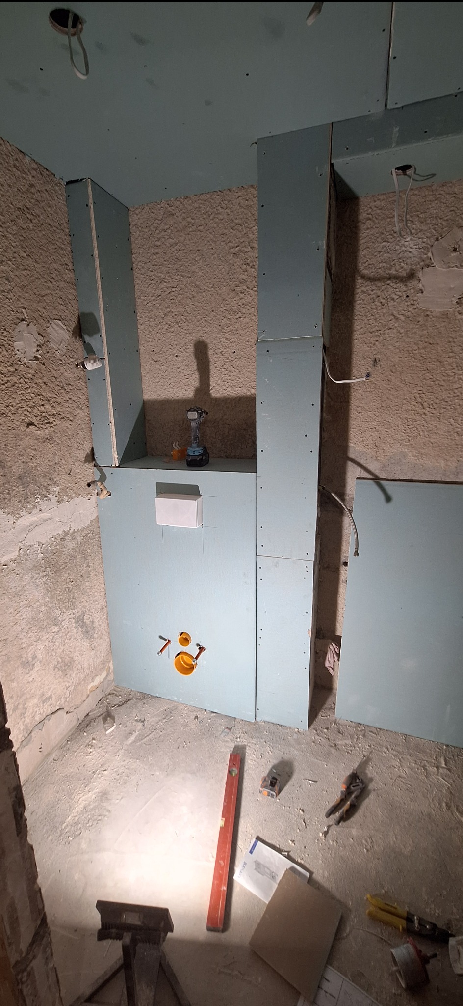 Montaż stelaża podtynkowego w łazience z widocznymi przyłączami wodno-kanalizacyjnymi. Na podłodze poziomica, narzędzia i dokumentacja. Ściany surowe, przed wykończeniem.