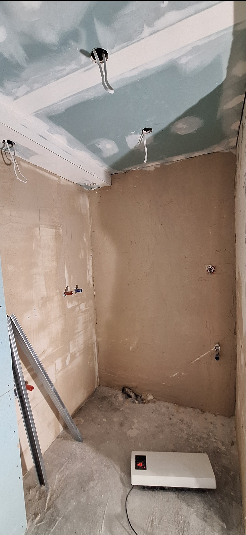 Surowe wnętrze łazienki w trakcie remontu: ściany po szpachlowaniu, sufit z płyt gipsowych z wystającymi kablami, grzejnik elektryczny na podłodze, profile aluminiowe.