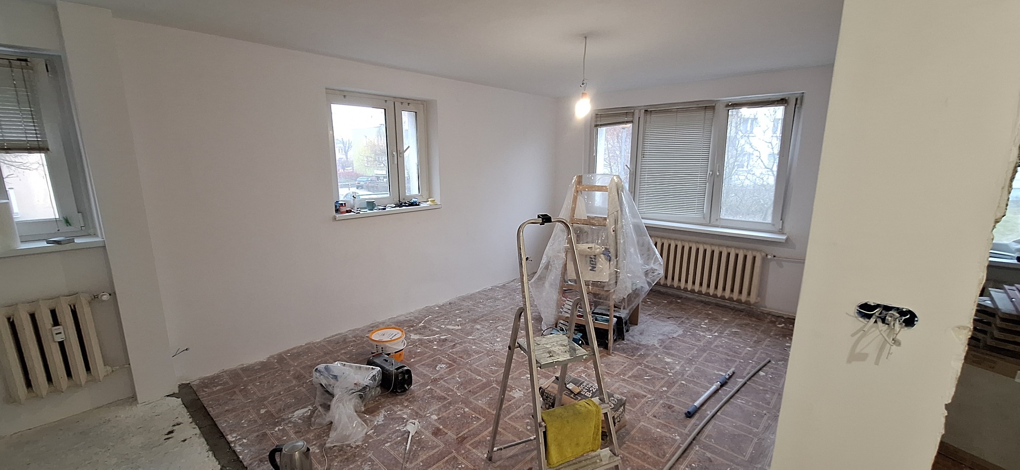 Pokój po malowaniu ścian na biało, widoczne narzędzia malarskie, drabina i zabezpieczone okno folią. Podłoga częściowo zakryta farbą. Gniazdko elektryczne bez osłony.