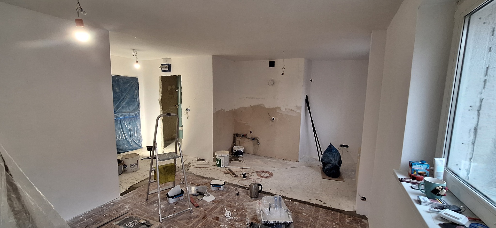 Wnętrze w trakcie remontu: ściany po szpachlowaniu i malowaniu, zabezpieczone drzwi, drabina, narzędzia malarskie, widoczne instalacje. Częściowo wykończone pomieszczenie.