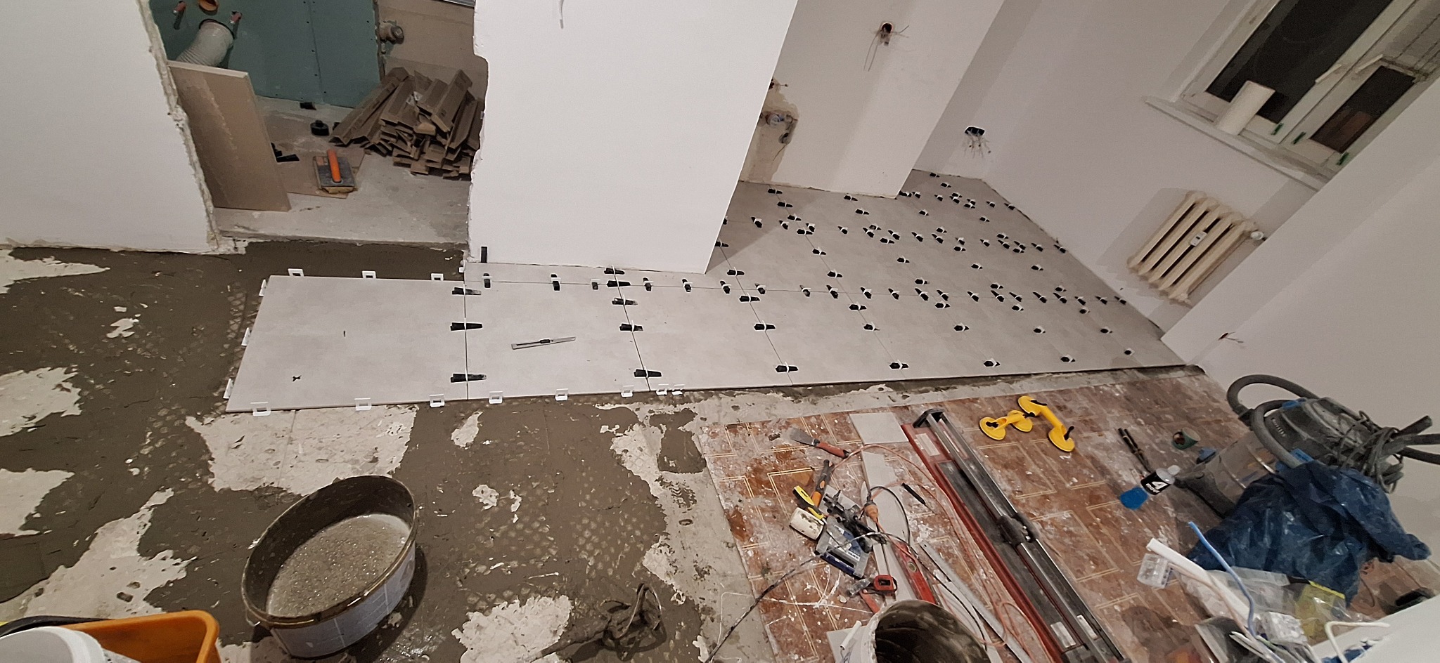Układanie jasnych płytek podłogowych z użyciem klipsów poziomujących. Widoczne narzędzia glazurnicze i wiadro z zaprawą. Fragment grzejnika przy oknie.