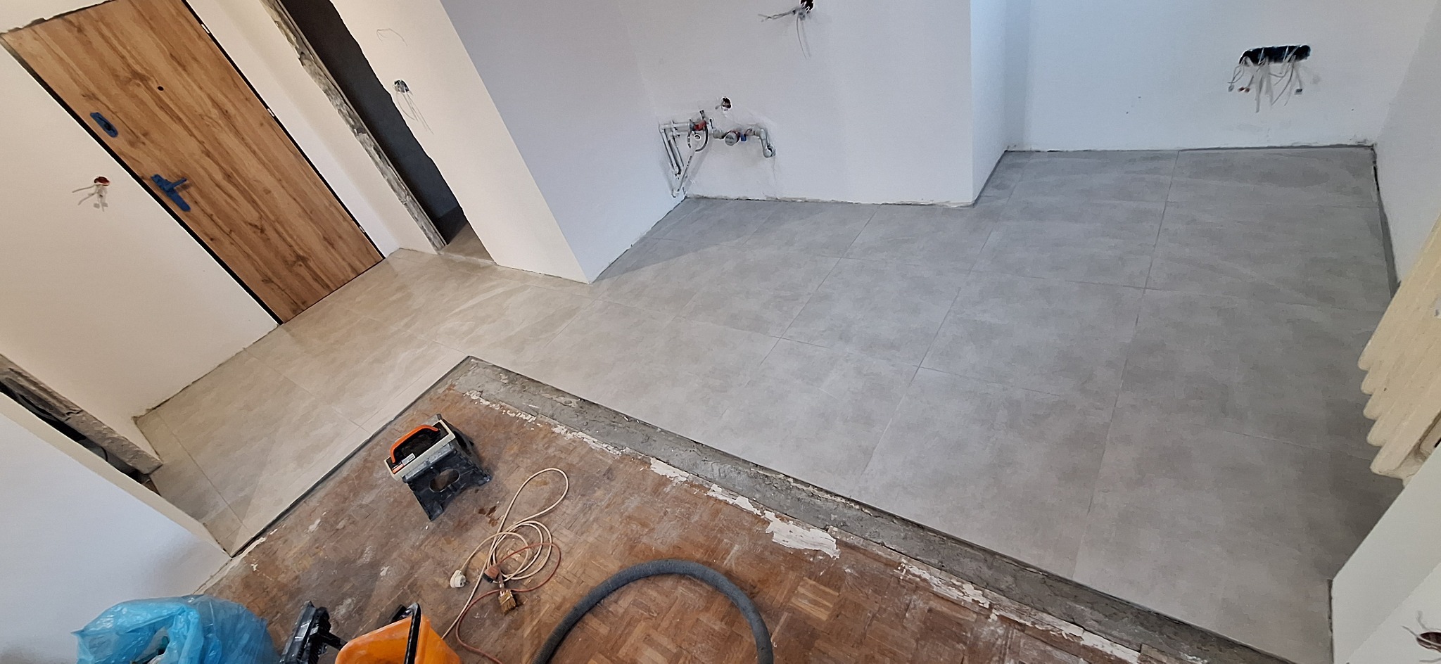 Ułożone szare płytki podłogowe w trakcie remontu łazienki, widoczne narzędzia i fragment starej podłogi. Ujęcie z góry, podkreślające postęp prac.