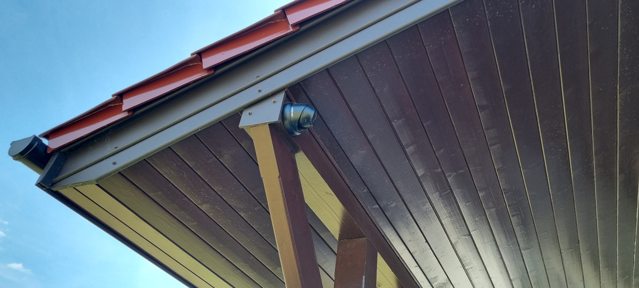 Kamera monitoringu kopułkowa zamontowana pod drewnianym zadaszeniem, widok z dołu na tle błękitnego nieba. Widoczne elementy konstrukcyjne dachu i orynnowanie.