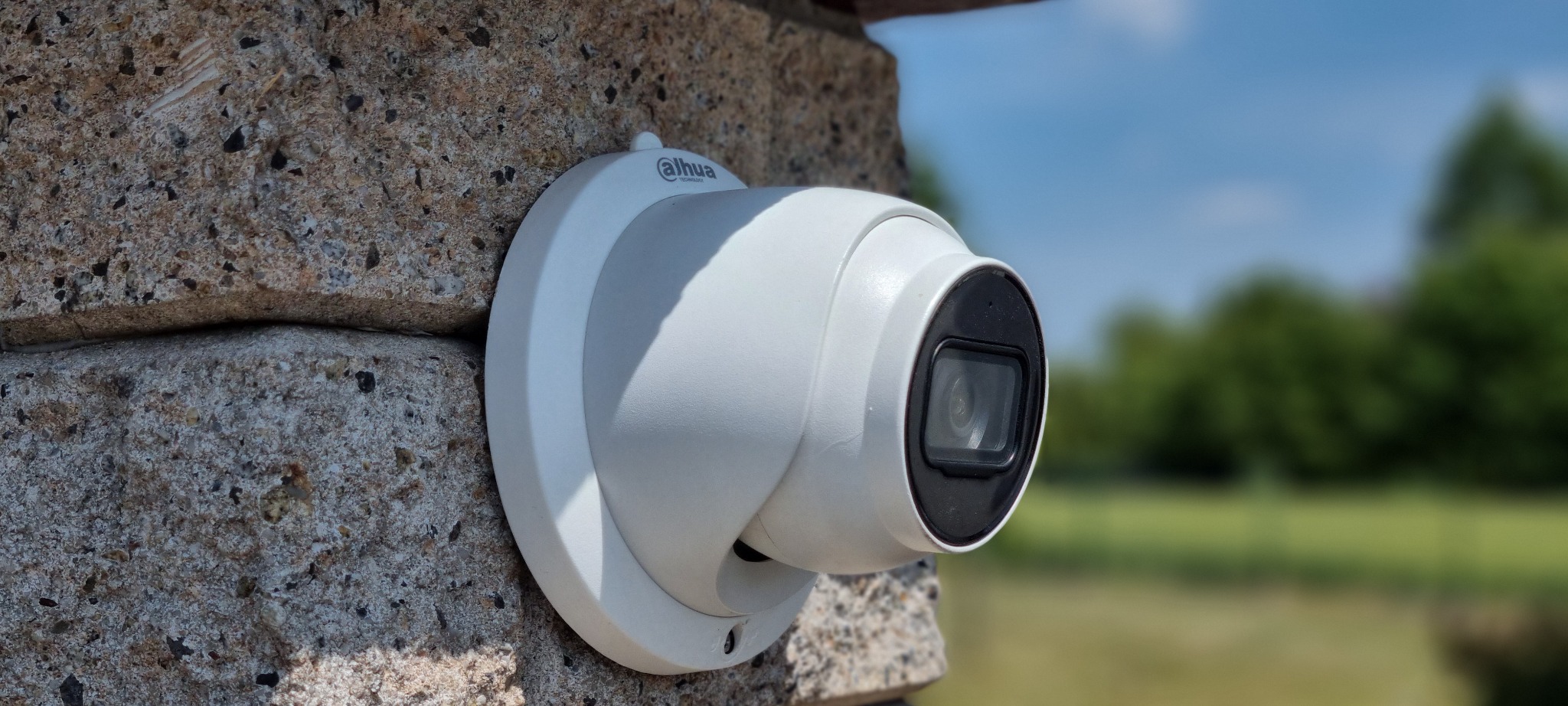 Kamera monitoringu Dahua zamontowana na szarej, kamiennej ścianie budynku. W tle rozmyte drzewa i błękitne niebo. Widok z boku, oświetlenie dzienne.