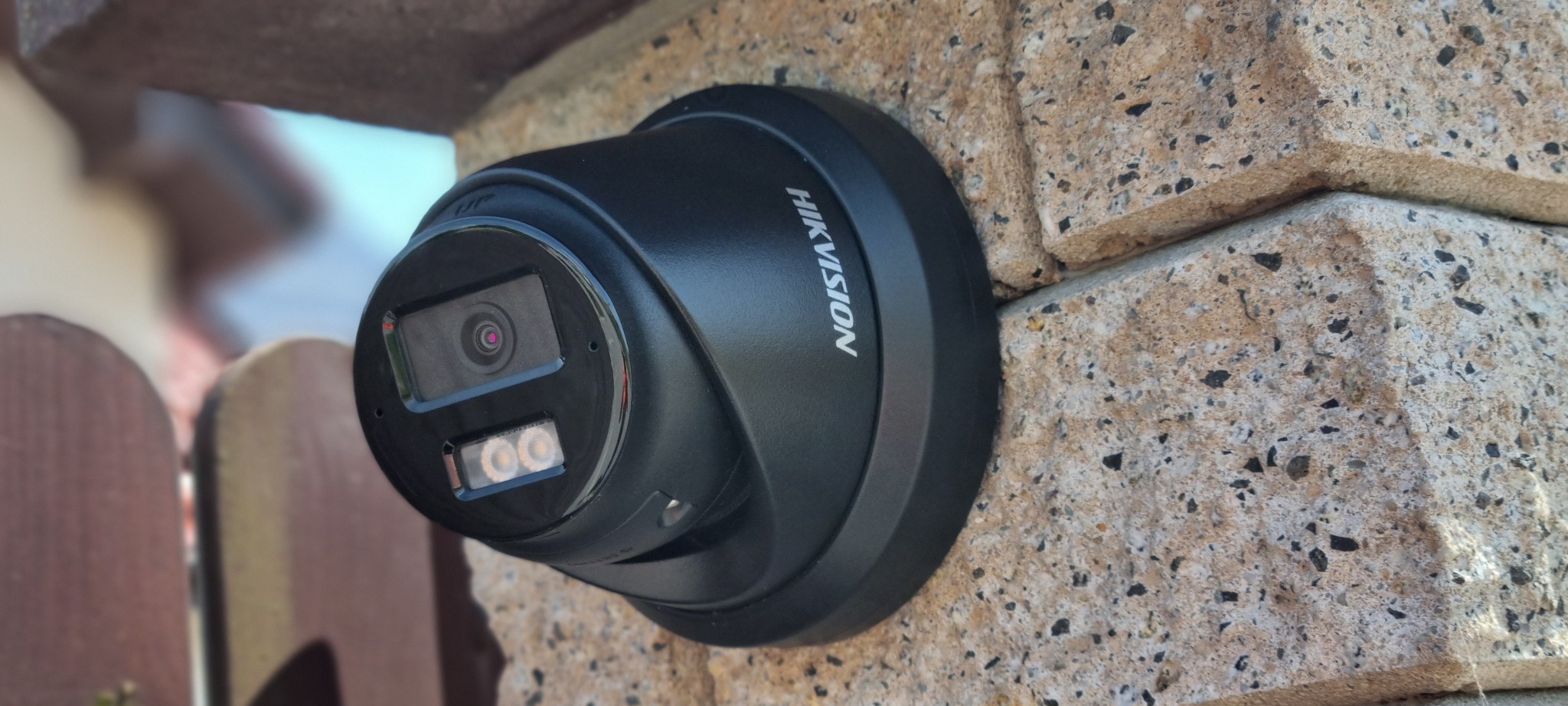 Kamera monitoringu Hikvision, kopułkowa, zamontowana na elewacji z kamienia dekoracyjnego. Widoczny obiektyw i diody IR. Częściowo widoczne ogrodzenie i fragment budynku.