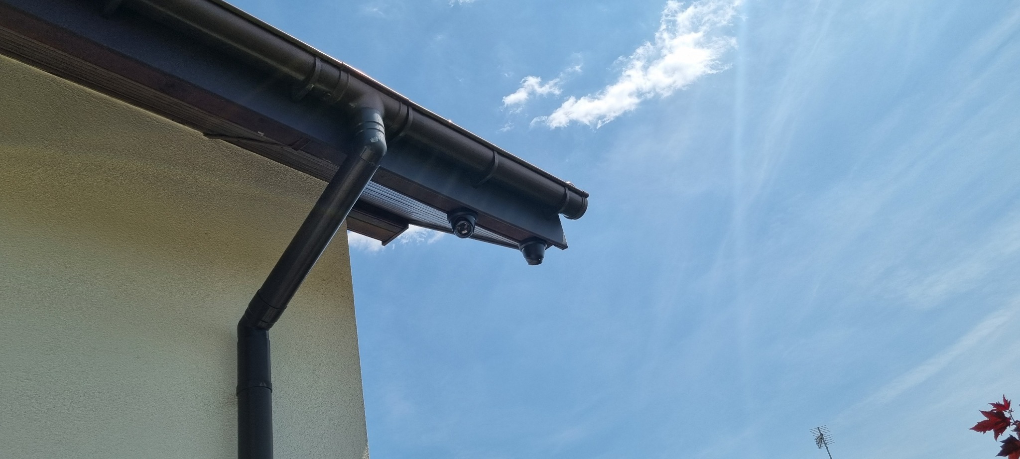Dwie kamery monitoringu kopułkowe zamontowane pod okapem budynku z brązowymi rynnami na tle błękitnego nieba z chmurami. Widok z dołu.