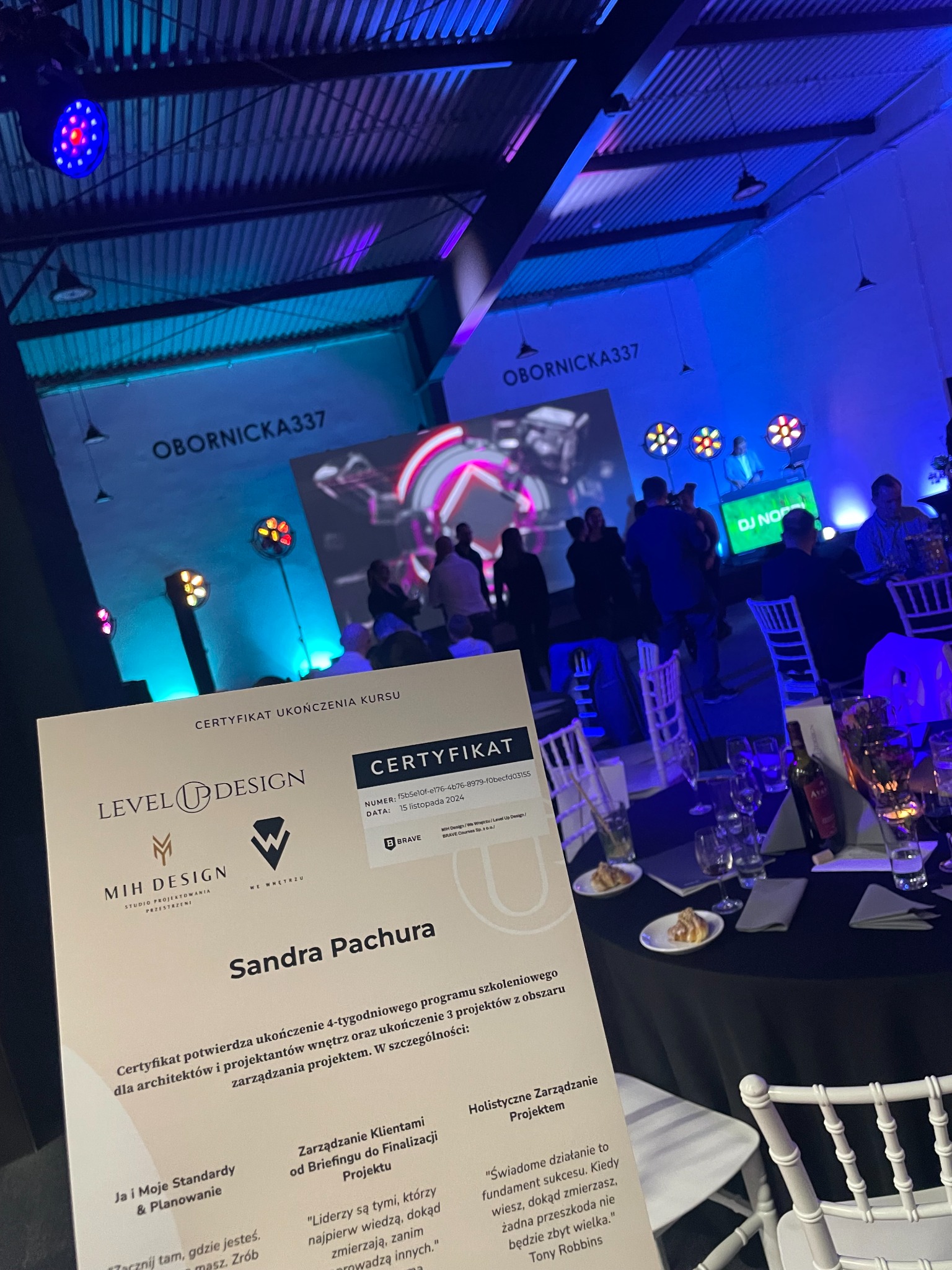 Certyfikat ukończenia kursu Level Up Design dla architektów wnętrz na tle sali eventowej z DJ-em i gośćmi. Dokument na stole, w tle scena i oświetlenie.