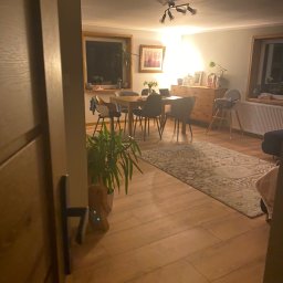 Smart Move Adrian Kulig - Jasne, przytulne wnętrze jadalni w Wiśle z drewnianą podłogą i stołem. Widok zza otwartych drzwi, z rośliną doniczkową w kadrze. Ciepłe oświetlenie.