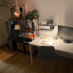 Smart Move Adrian Kulig - Nowoczesne biurko z laptopem, lampką i monitorem, ustawione przy ścianie z półkami. Na półkach rośliny doniczkowe i dekoracje. Minimalistyczny styl, ciepłe oświetlenie.