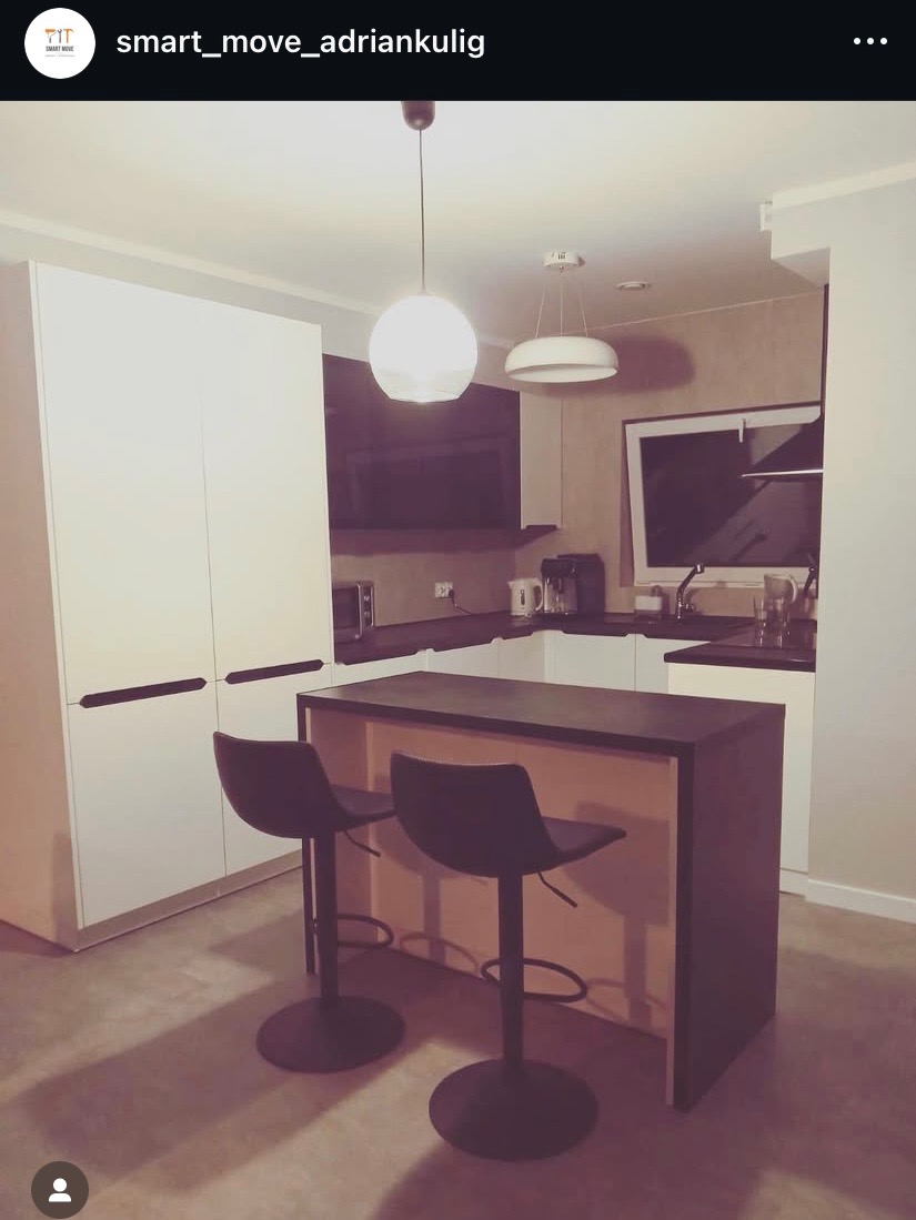 Nowoczesna kuchnia z wyspą i dwoma hokerami. Białe szafki, ciemny blat, minimalistyczne oświetlenie. Ujęcie z niskiego kąta, podkreślające przestrzeń.