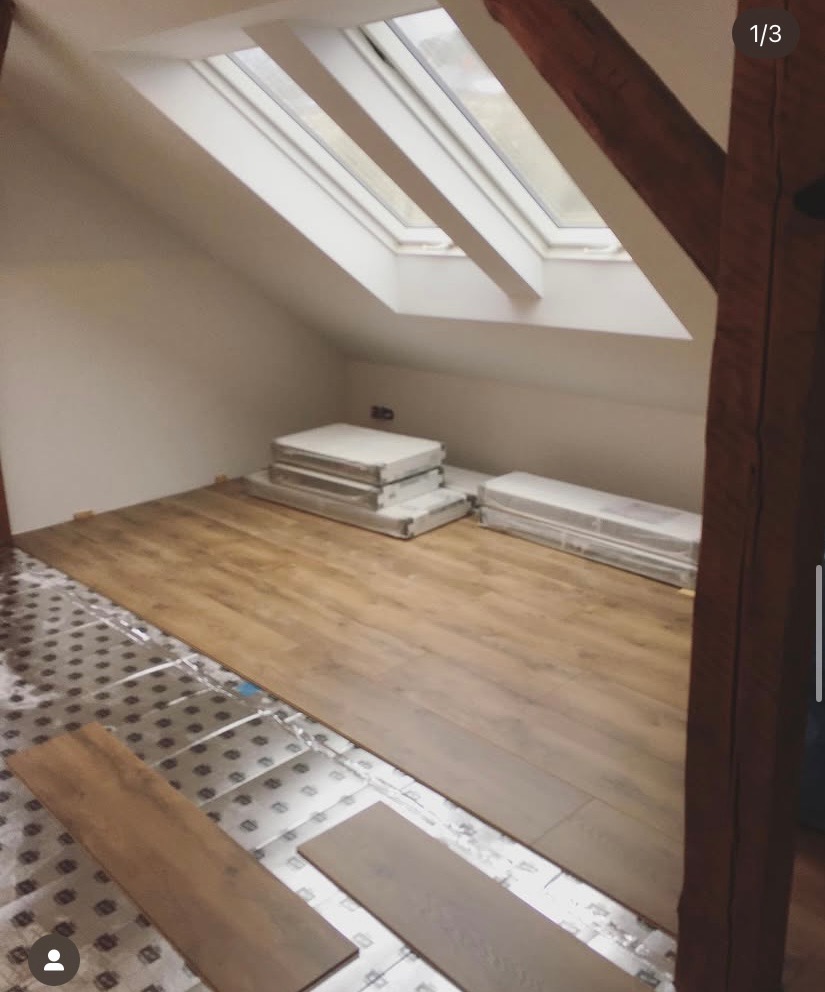 Montaż paneli podłogowych w pokoju na poddaszu z oknem dachowym. Częściowo ułożona podłoga, widoczne materiały izolacyjne i panele do ułożenia. Drewniane elementy konstrukcyjne.