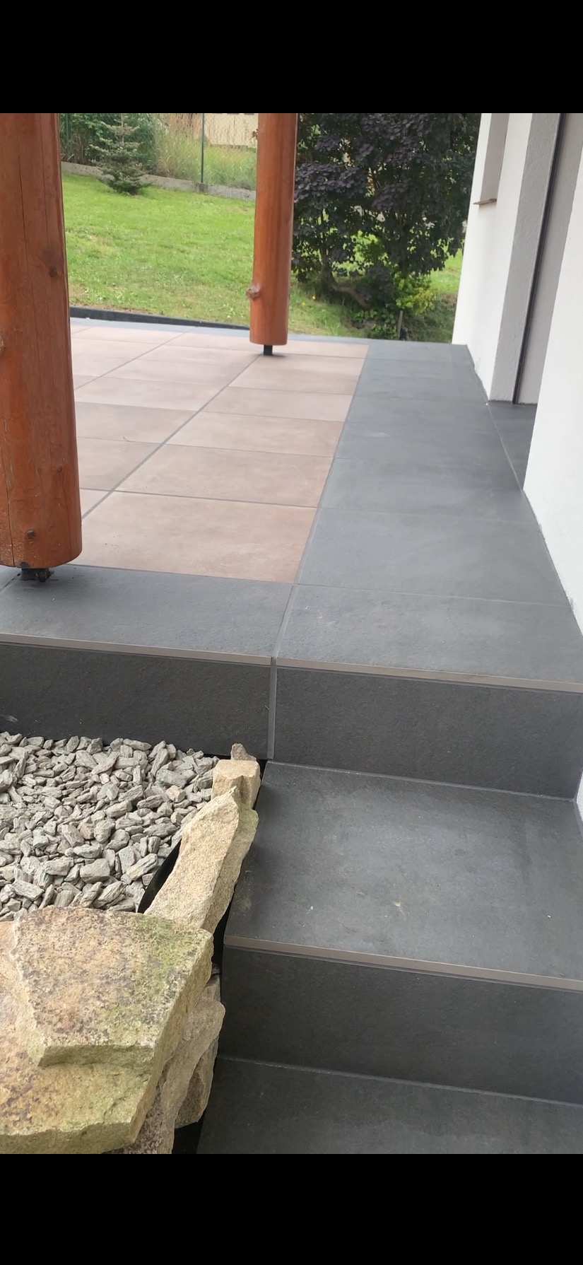 Wykończony taras z szarymi płytkami schodowymi i jasnymi płytami na podłodze. Widoczne drewniane słupy i kamienna rabata. Estetyczne wykończenie zewnętrzne.