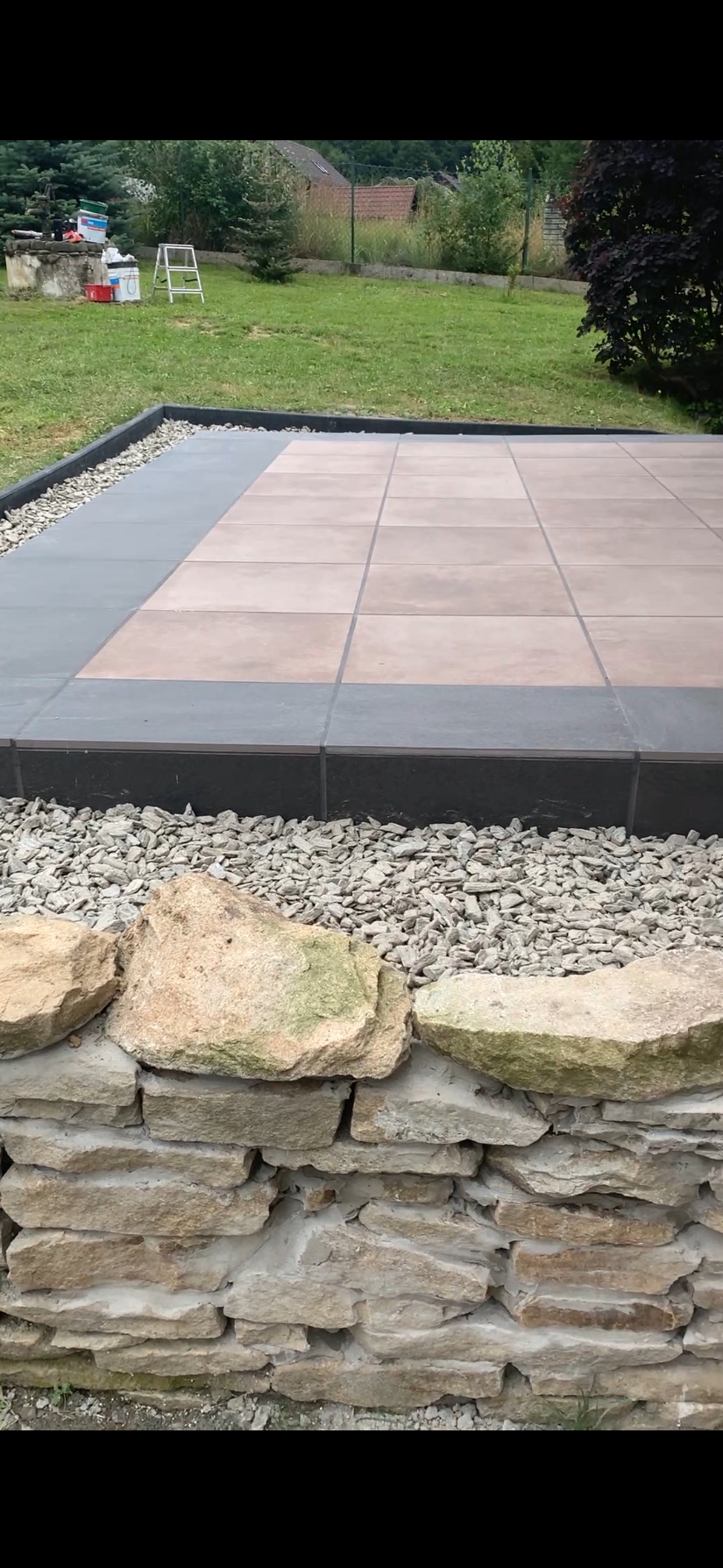 Wykończony taras z kamienną obudową i jasnymi płytkami, otoczony zielenią. Widoczne drobne kamienie dekoracyjne. Ujęcie z bliska, podkreślające detale wykończenia.