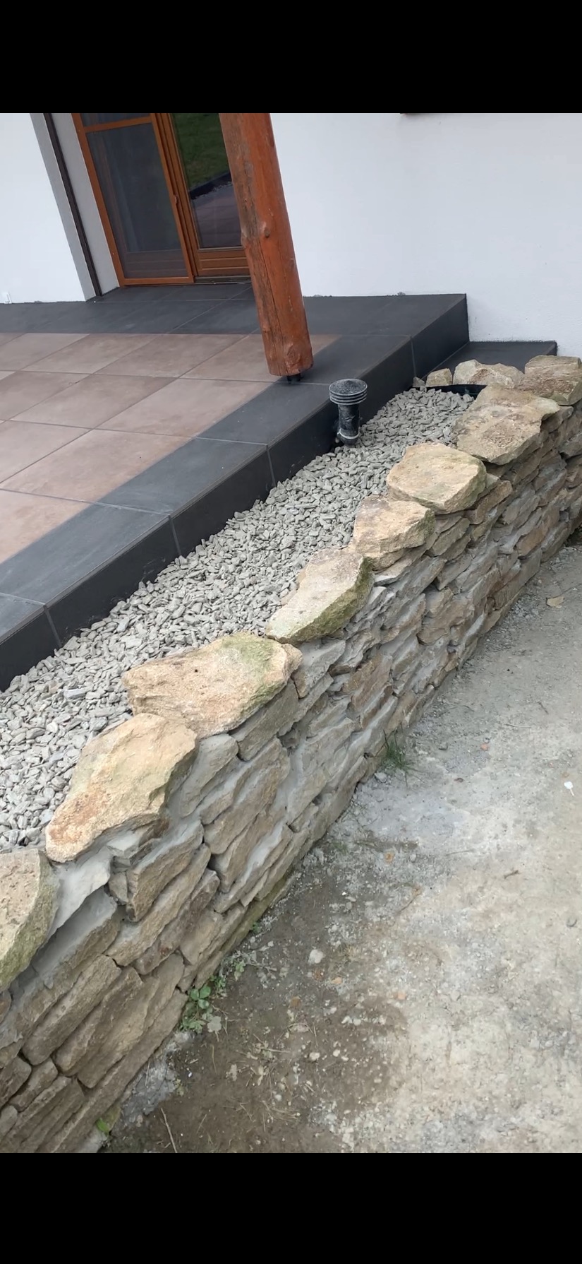 Kamienny murek oddzielający taras z brązowych płytek od szarego podłoża. W tle biała ściana budynku z drewnianymi drzwiami i elementem drewnianej konstrukcji.