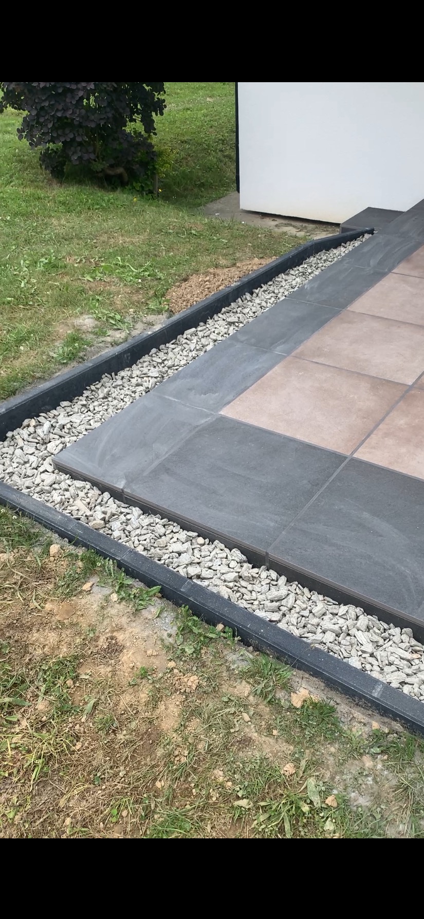 Ułożona, kwadratowa, brązowo-szara płytka tarasowa z obramowaniem z czarnego tworzywa i białych kamyków na trawniku przy budynku.