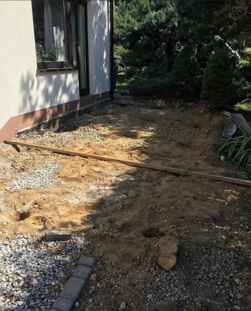Przygotowanie terenu pod taras: wykopane dołki, rozsypany żwir, listwa poziomująca przy ścianie domu. Widok na ogród w tle.