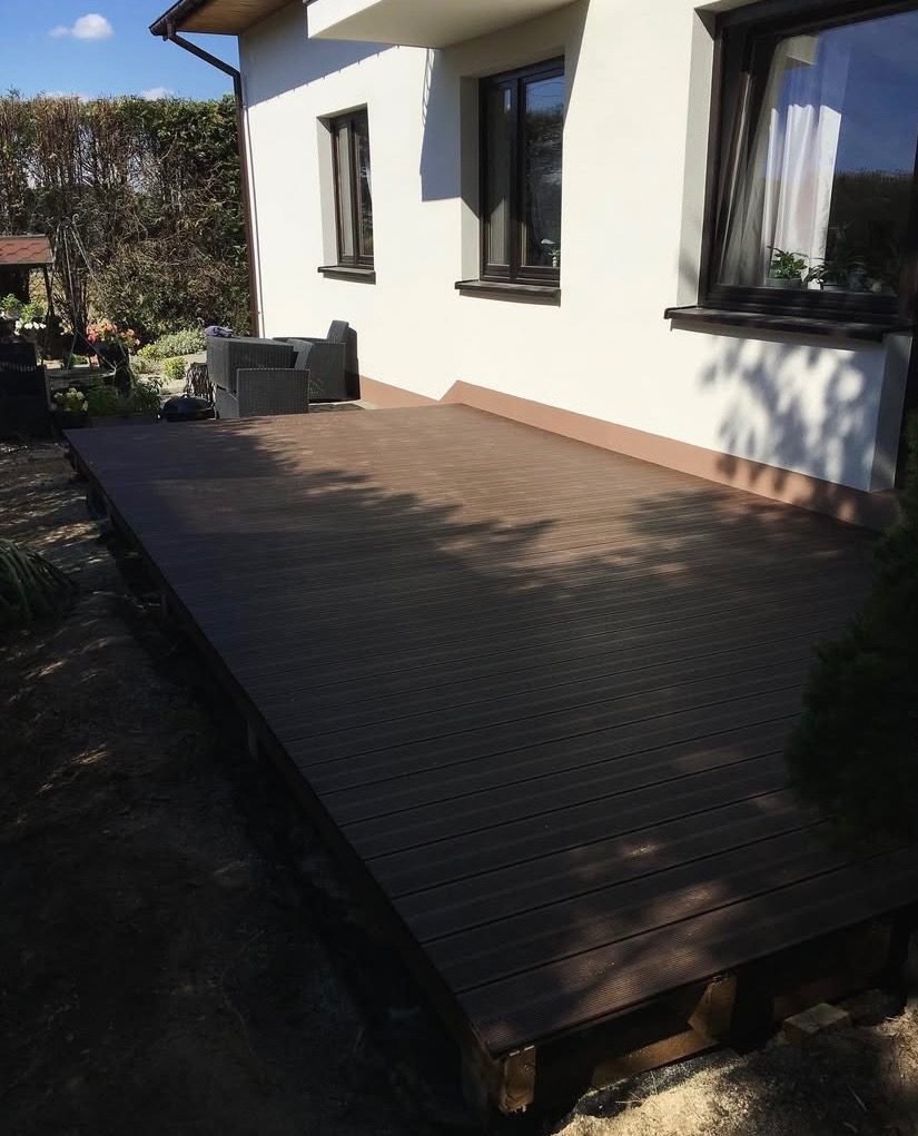 Nowoczesny taras z desek kompozytowych przylegający do białego domu z ciemnymi oknami. Widoczne meble ogrodowe i zieleń. Cień drzew na desce.