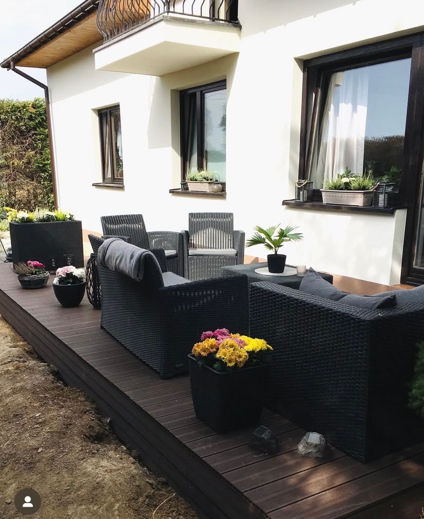 Nowoczesny taras z ciemnego drewna kompozytowego przy białym domu. Czarne meble ogrodowe z poduszkami i donice z kwiatami. Elegancki design.