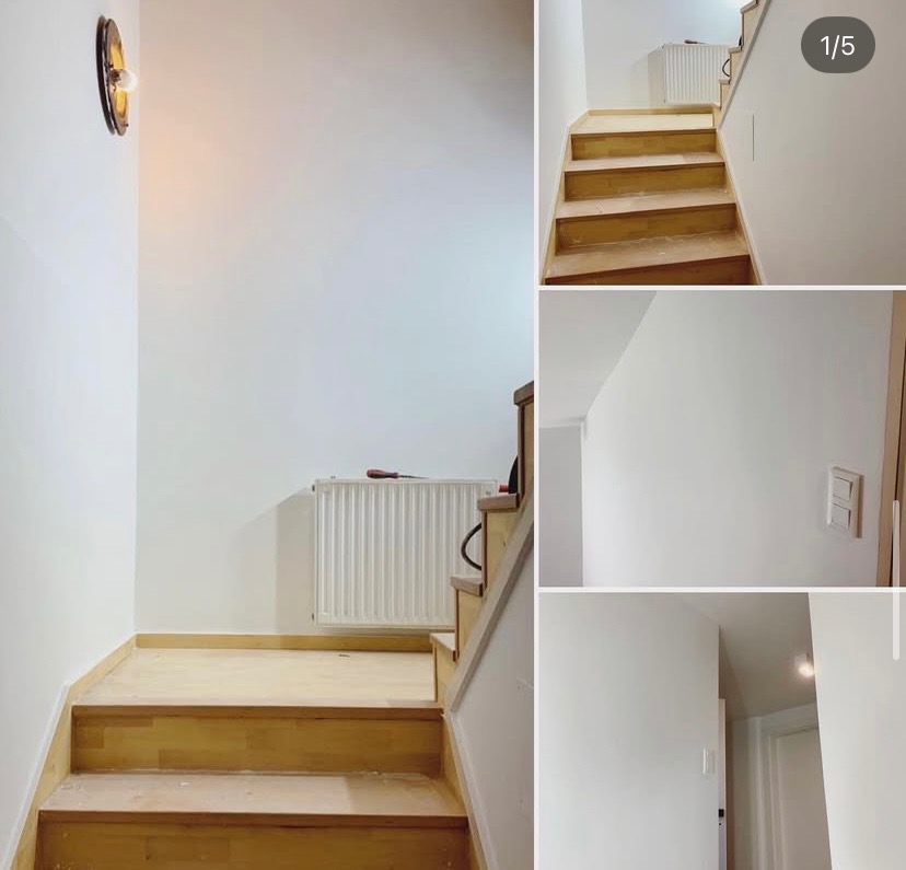 Klatka schodowa po malowaniu na biało, widoczne drewniane schody, grzejnik i lampa ścienna. Ujęcie z dołu schodów, jasne wnętrze.