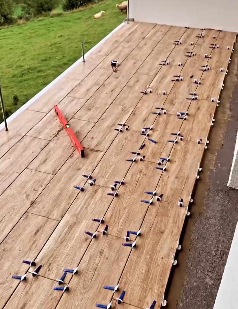 Układanie drewnopodobnych płytek na tarasie z użyciem poziomicy i klipsów poziomujących. W tle trawnik i pasące się owce.