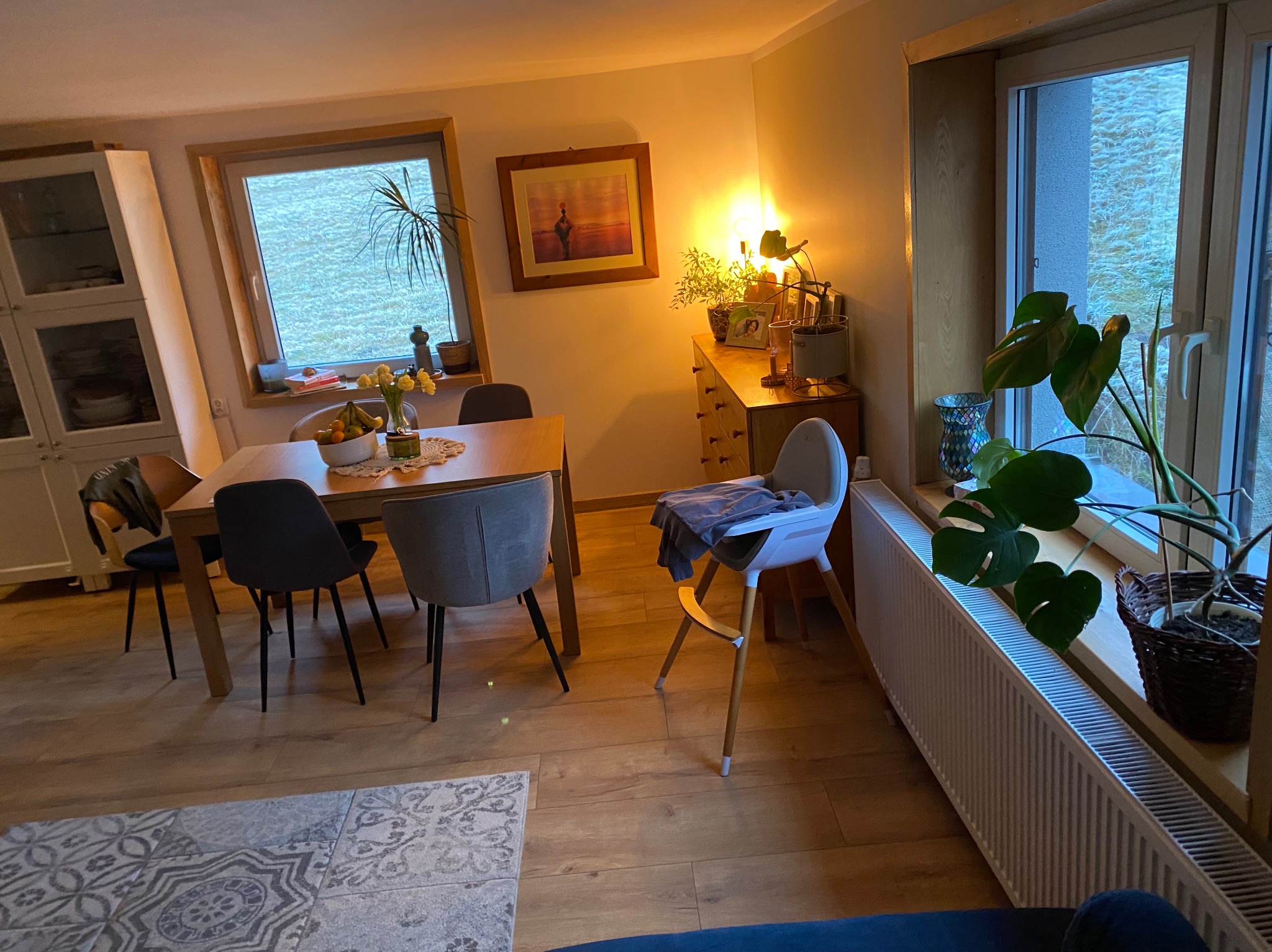 Jasne wnętrze jadalni z drewnianą podłogą i stołem. Widoczne krzesła, okno z roślinami i fragment dekoracyjnej podłogi. Ciepłe oświetlenie.