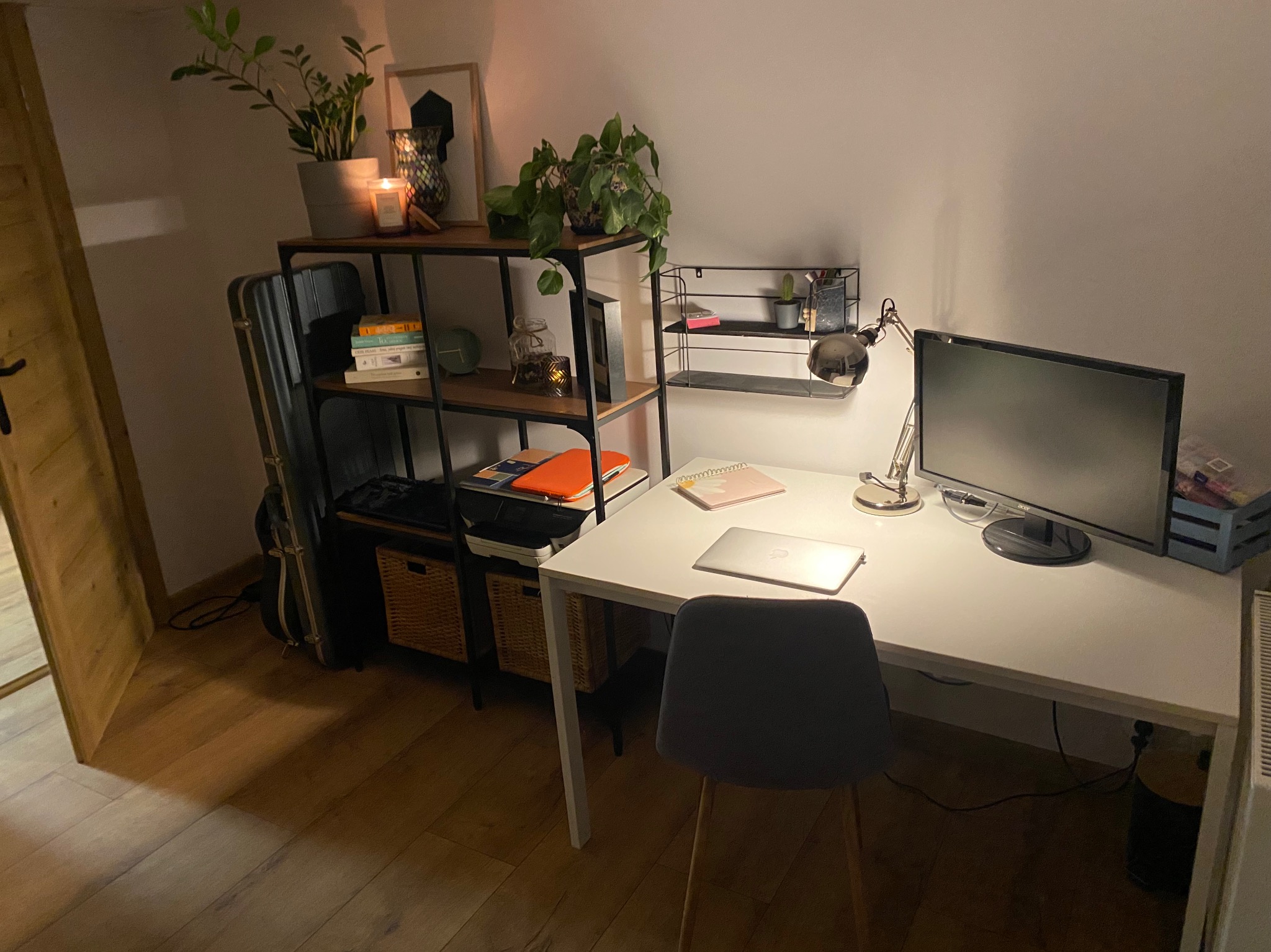 Nowoczesne biurko z laptopem, lampką i monitorem, ustawione przy ścianie z półkami. Na półkach rośliny doniczkowe i dekoracje. Minimalistyczny styl, ciepłe oświetlenie.