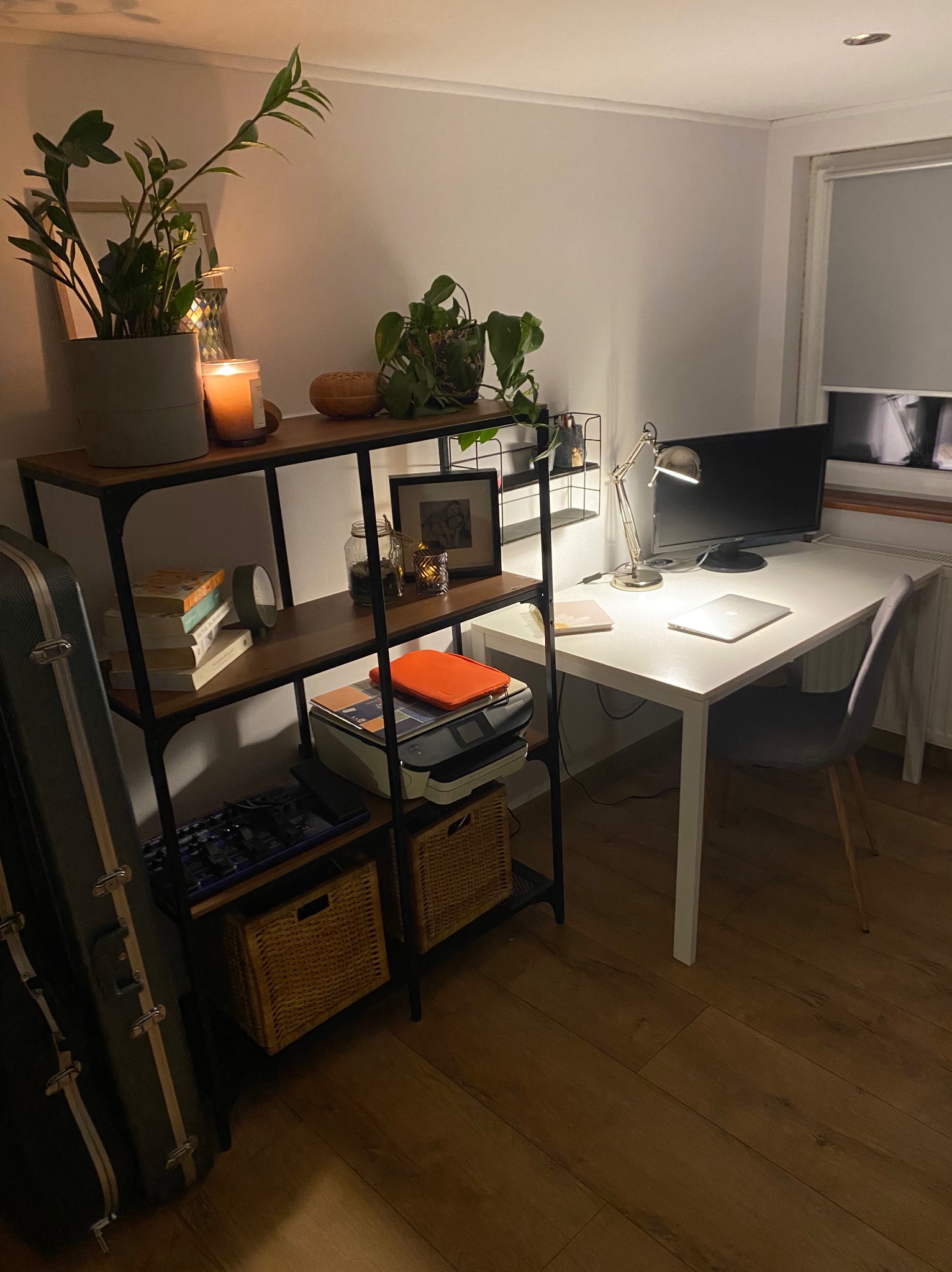 Domowe biuro z białym biurkiem, lampką, komputerem i regałem z roślinami, książkami i dekoracjami. Ciepłe oświetlenie tworzy przytulną atmosferę w pokoju.