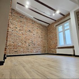 MMK INVEST GROUP SPÓŁKA Z OGRANICZONĄ ODPOWIEDZIALNOŚCIĄ - Wykończone wnętrze z ceglaną ścianą, drewnianą podłogą i białym oknem. Widoczne elementy dekoracyjne sufitu i grzejnik. Minimalistyczny styl wykończenia.