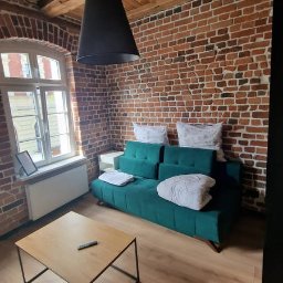 MMK INVEST GROUP SPÓŁKA Z OGRANICZONĄ ODPOWIEDZIALNOŚCIĄ - Loftowe wnętrze z ceglaną ścianą, drewnianym sufitem i turkusową sofą. Na podłodze panele, a na stoliku pilot. Widoczne okno z białymi ramami i czarna lampa.