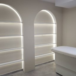 MMK INVEST GROUP SPÓŁKA Z OGRANICZONĄ ODPOWIEDZIALNOŚCIĄ - Minimalistyczna łazienka z podświetlanymi wnękami w kształcie łuku i nowoczesnym fotelem zabiegowym. Szare płytki podłogowe i stonowana kolorystyka ścian.