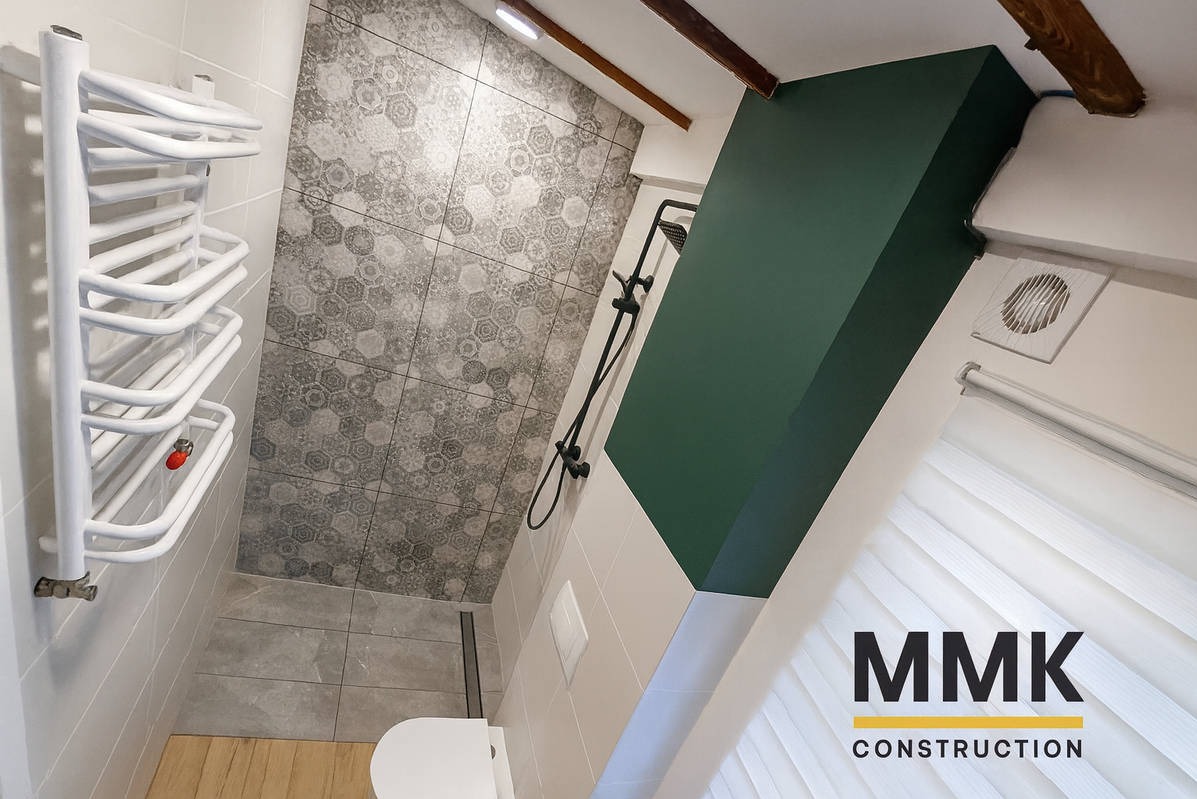 Nowoczesna łazienka na poddaszu z geometrycznymi płytkami, czarną armaturą i białym grzejnikiem drabinkowym. Ukośna ściana pomalowana na butelkową zieleń.