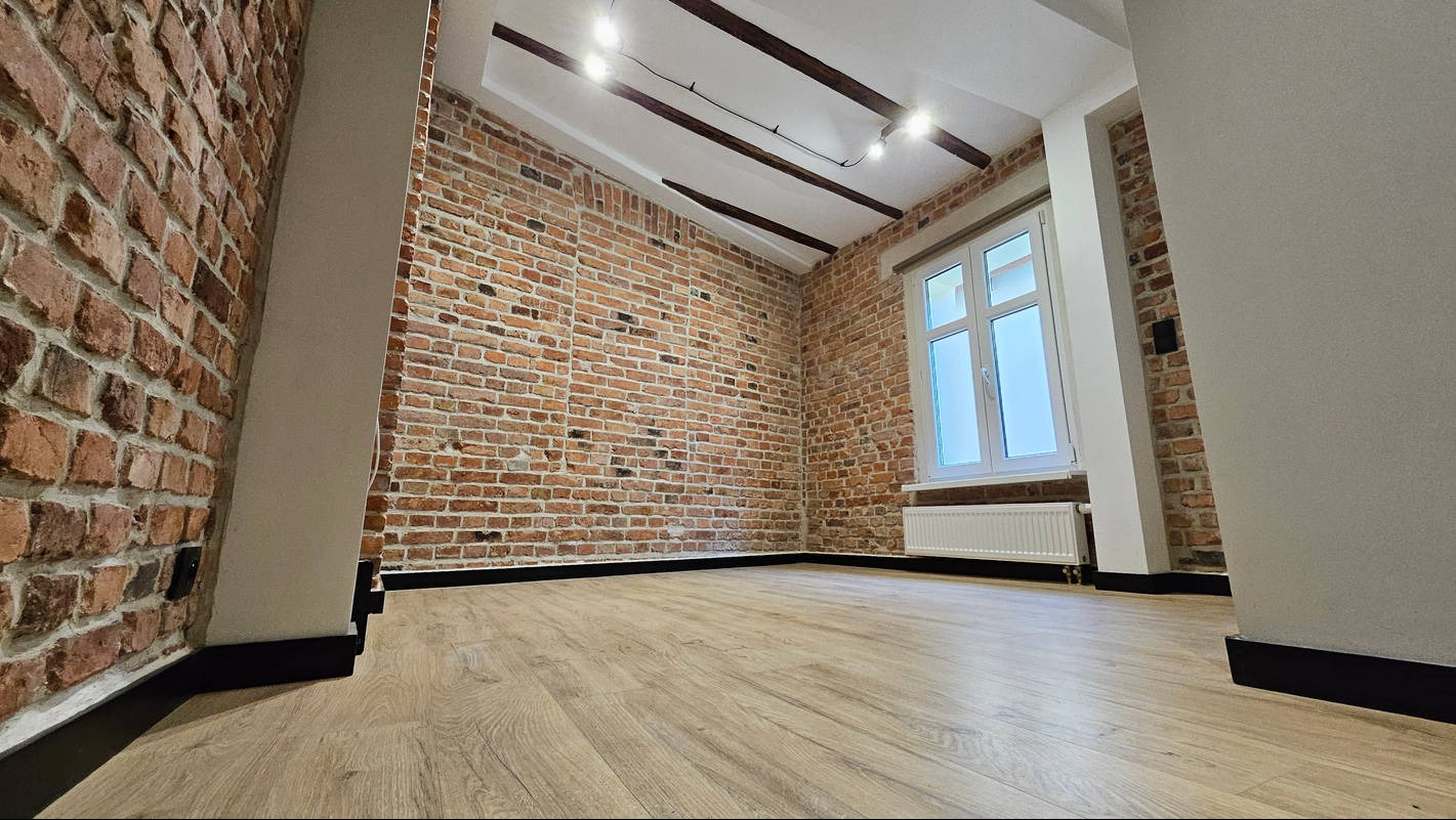 Wykończone wnętrze z ceglaną ścianą, drewnianą podłogą i białym oknem. Widoczne elementy dekoracyjne sufitu i grzejnik. Minimalistyczny styl wykończenia.