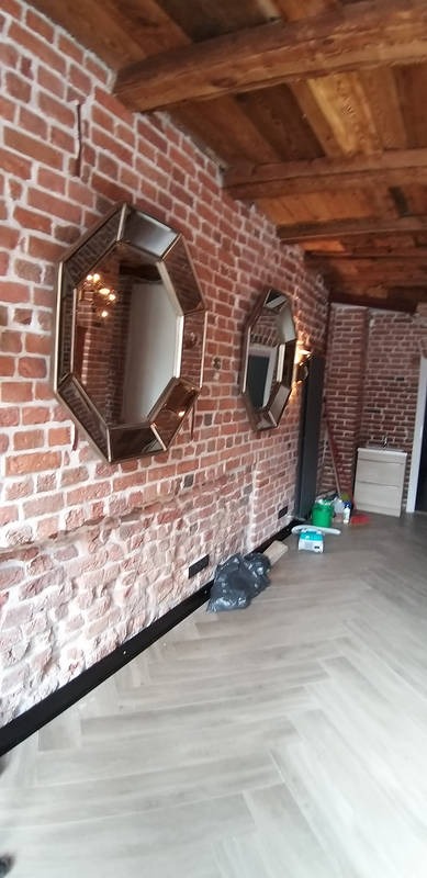 Wykończone wnętrze z ceglaną ścianą, drewnianym sufitem i parkietem w jodełkę. Dwa ośmiokątne lustra z metalową ramą na ścianie. Widoczne akcesoria i narzędzia.