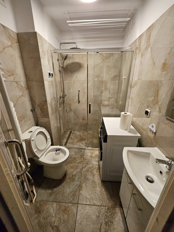 Wykończona łazienka z kabiną prysznicową, umywalką i toaletą w beżowych płytkach imitujących marmur. Widoczna pralka i suszarka podsufitowa. Ujęcie z perspektywy wejścia.