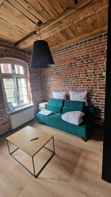 Loftowe wnętrze z ceglaną ścianą, drewnianym sufitem i turkusową sofą. Na podłodze panele, a na stoliku pilot. Widoczne okno z białymi ramami i czarna lampa.