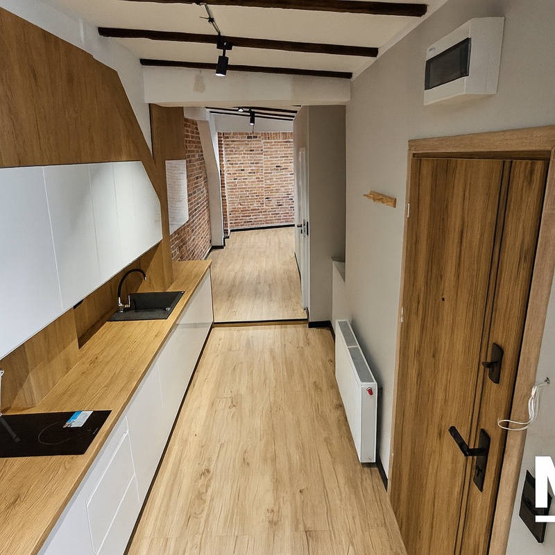Nowoczesna kuchnia z drewnianym blatem i białymi szafkami, widok na korytarz z ceglaną ścianą. Oświetlenie punktowe i drewniane belki stropowe dodają charakteru.