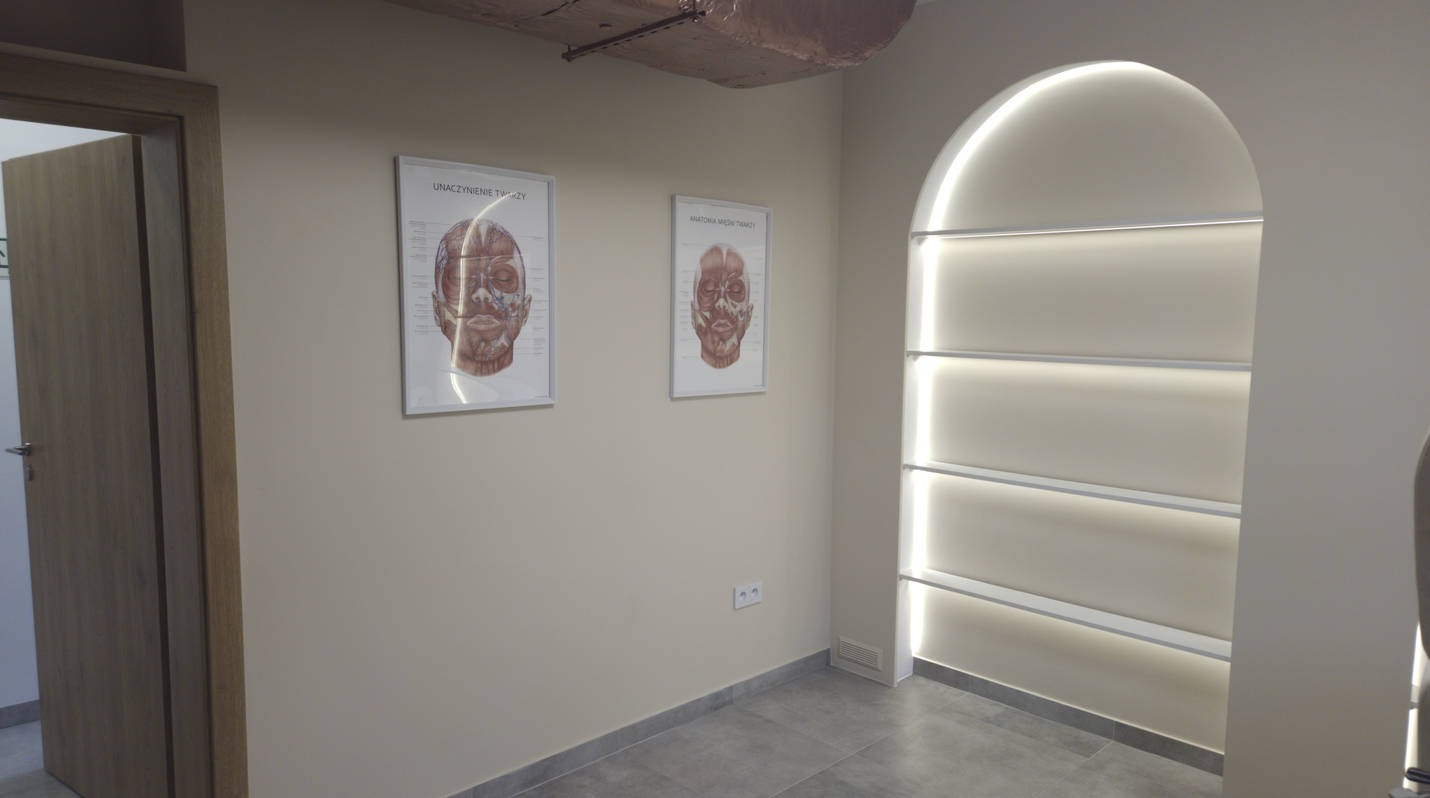 Jasne wnętrze z oświetloną wnęką w kształcie łuku i dwoma plakatami anatomicznymi na ścianie. Minimalistyczny design, szare płytki podłogowe.