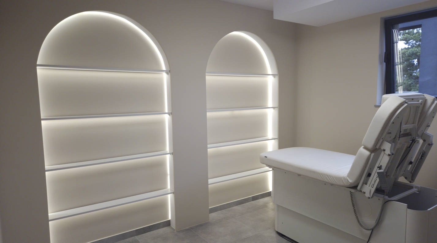 Minimalistyczna łazienka z podświetlanymi wnękami w kształcie łuku i nowoczesnym fotelem zabiegowym. Szare płytki podłogowe i stonowana kolorystyka ścian.