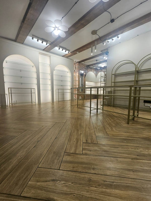 Jasne wnętrze sklepu z drewnopodobną podłogą ułożoną w jodełkę, białymi ścianami z wnękami i drewnianymi belkami stropowymi. Metalowe regały w złotym kolorze.