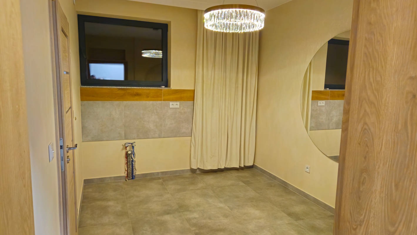 Wnętrze łazienki w trakcie prac: położone płytki pod oknem, widoczne wystające rury, okrągłe lustro na ścianie i nowoczesny żyrandol. Neutralna kolorystyka.