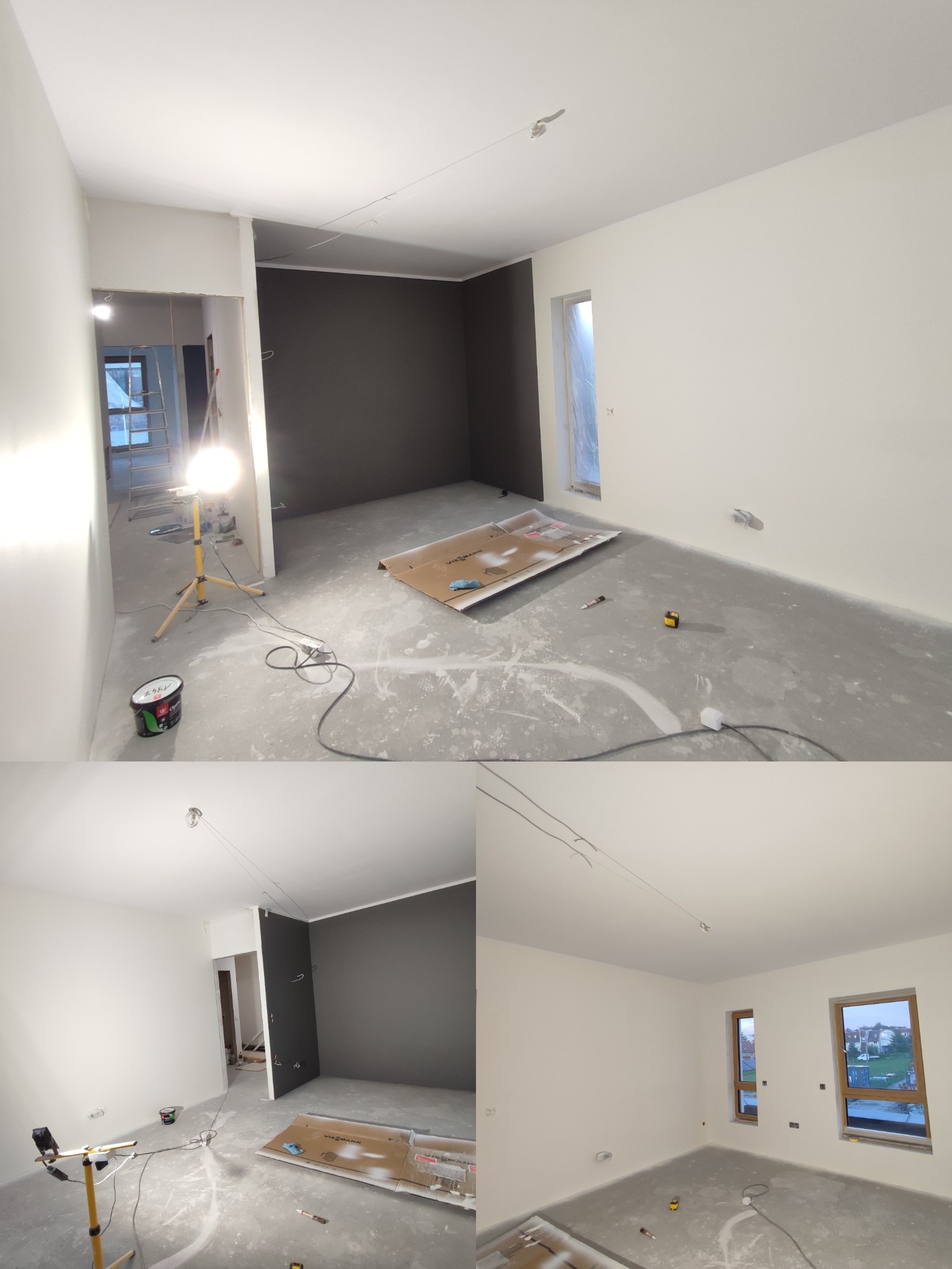 Wnętrze po malowaniu: białe ściany, jedna ściana grafitowa, widoczne narzędzia malarskie i lampa budowlana. Podłoga zabezpieczona kartonem. Widok na sąsiednie pomieszczenie.