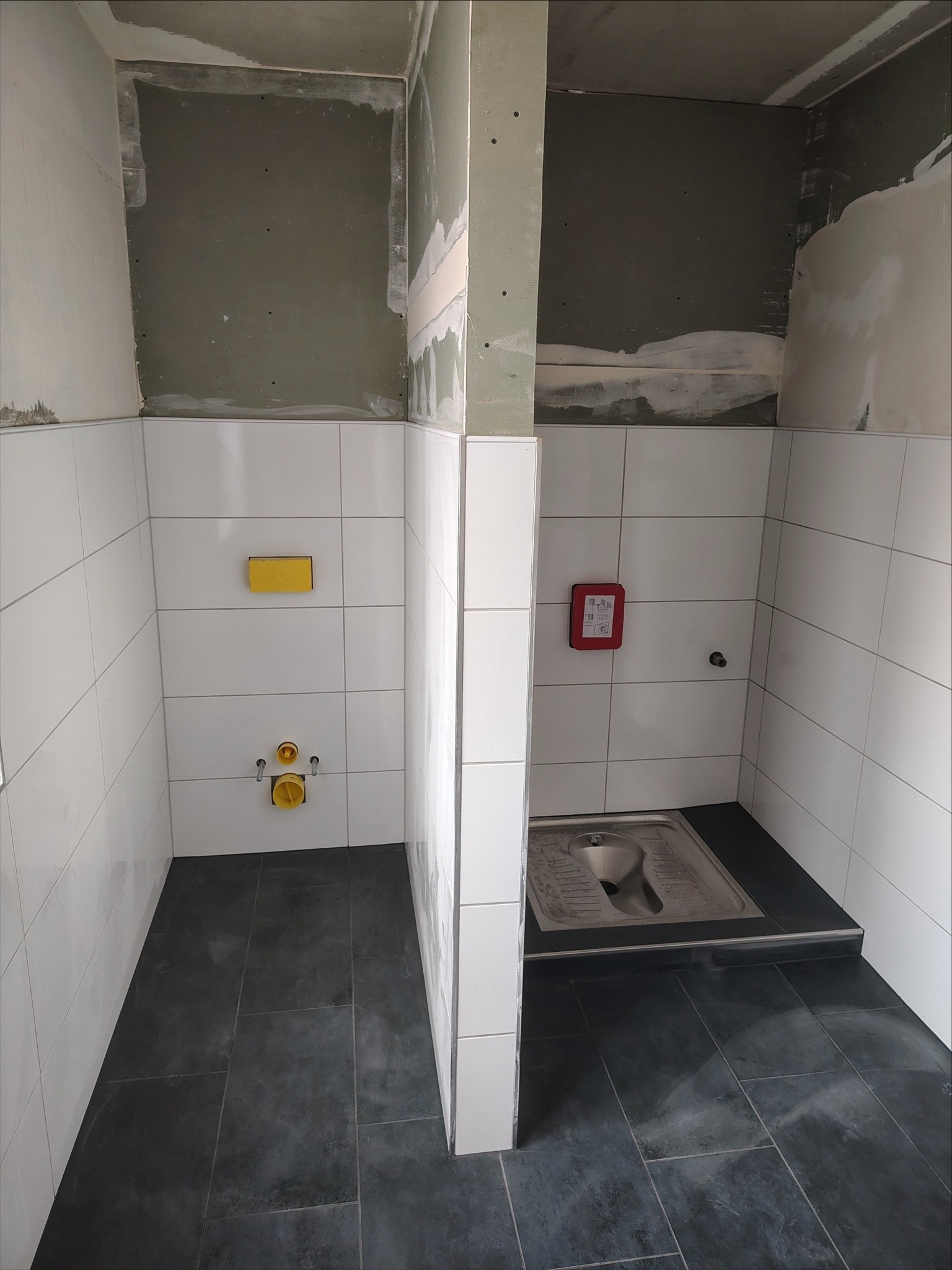 Wykończona kabina WC z białą glazurą ścienną i ciemną podłogą w Kowala Stępocina. Widoczne przyłącza i częściowo wykończone ściany działowe.