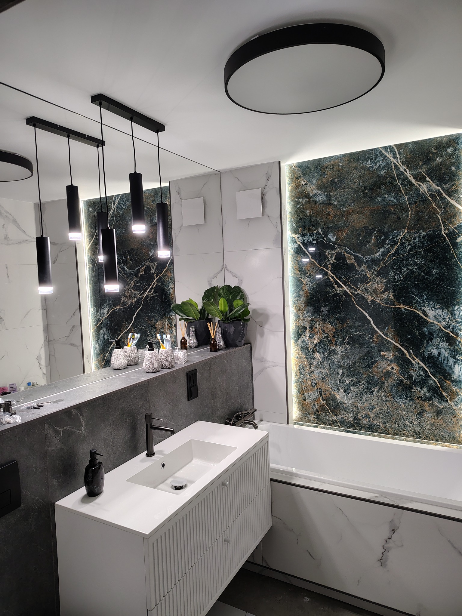 Nowoczesna łazienka z marmurowymi płytkami, podświetlanym panelem dekoracyjnym i wiszącą szafką umywalkową z czarną armaturą. Minimalistyczne oświetlenie.