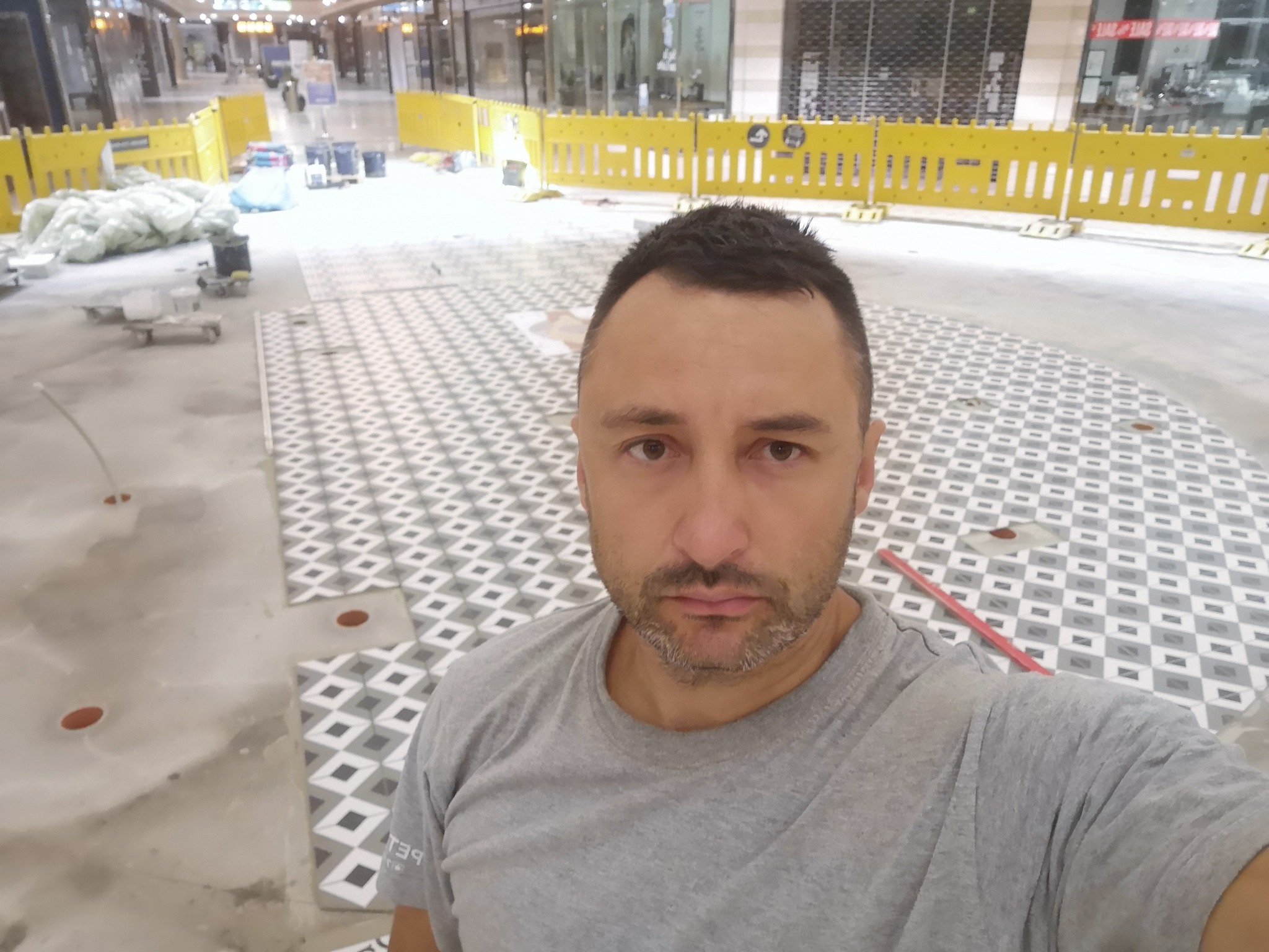 Układanie wzorzystych płytek podłogowych w centrum handlowym, widoczne żółte barierki zabezpieczające teren prac. Selfie osoby nadzorującej postęp prac.