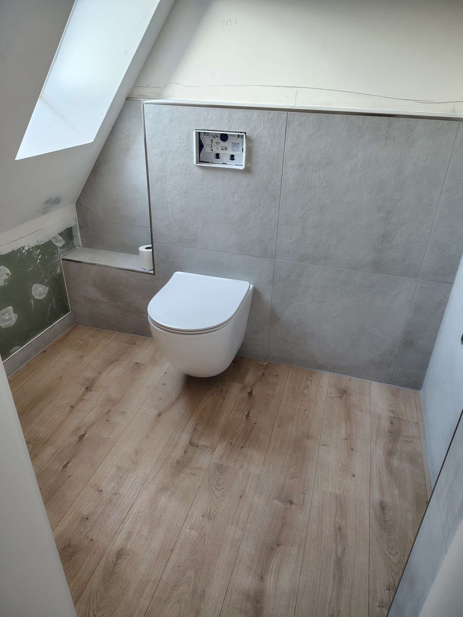 Wykończona łazienka na poddaszu z drewnopodobną podłogą i szarą glazurą ścienną. Widoczna podtynkowa toaleta i fragment ściany z niedokończonymi pracami wykończeniowymi.