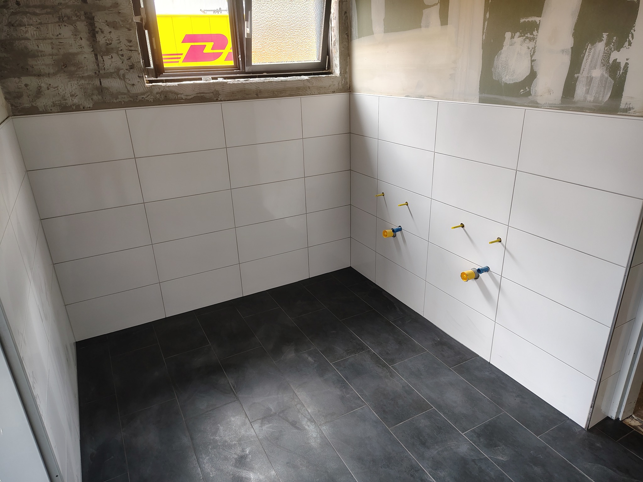 Wykończona łazienka z białymi płytkami ściennymi i ciemną podłogą. Widoczne przyłącza wodne na ścianie. W oknie odbija się żółty samochód dostawczy.