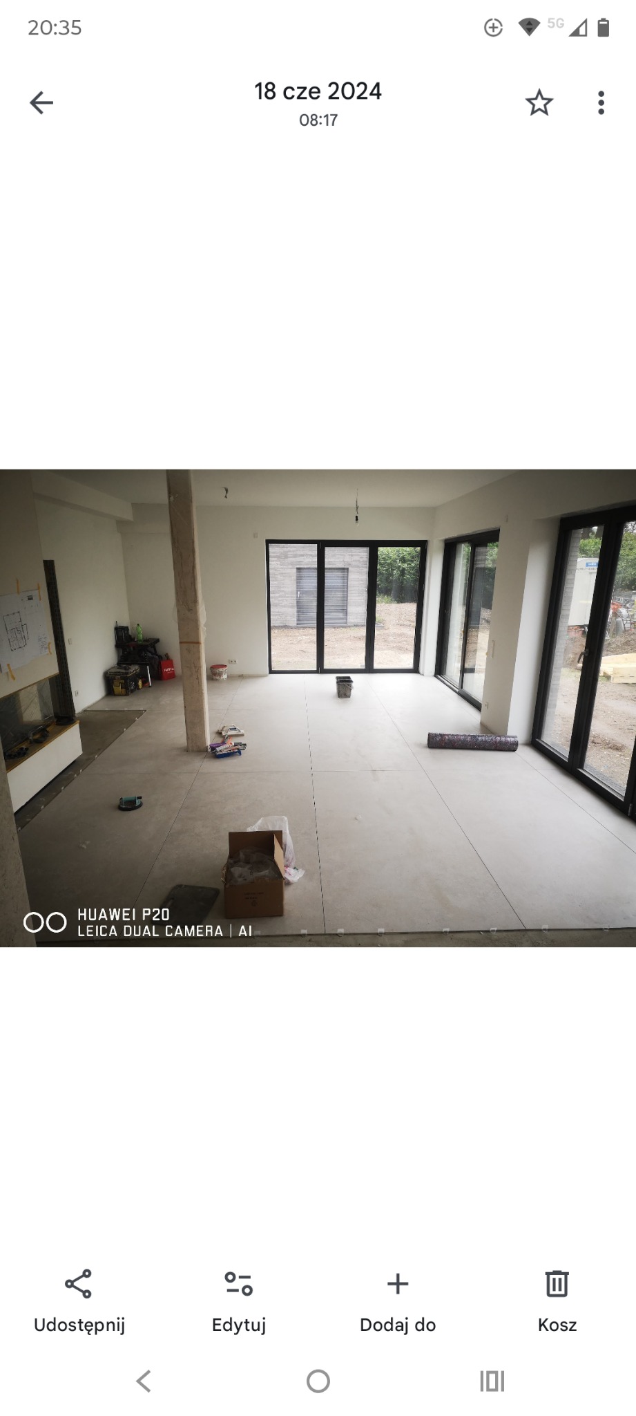 Jasne wnętrze z ułożoną, dużą, szarą terakotą. Widoczne narzędzia i karton. Duże okna przesuwne w czarnych ramach. Minimalistyczny styl wykończenia.