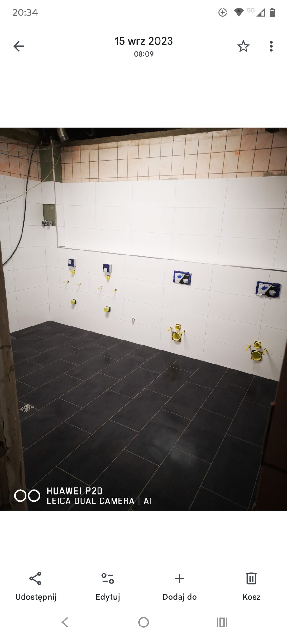 Wykończona łazienka z czarną podłogą z płytek i białymi płytkami na ścianach. Widoczne elementy instalacji sanitarnej. Minimalistyczny design.